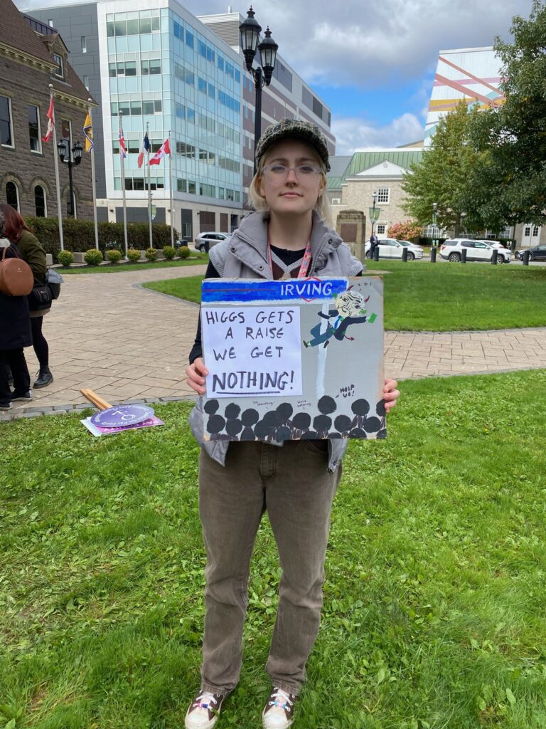 image of young person in front of the Fredericton Legislative Building holding a sign that says "Higgs Gets a Raise, We Get Nothing"