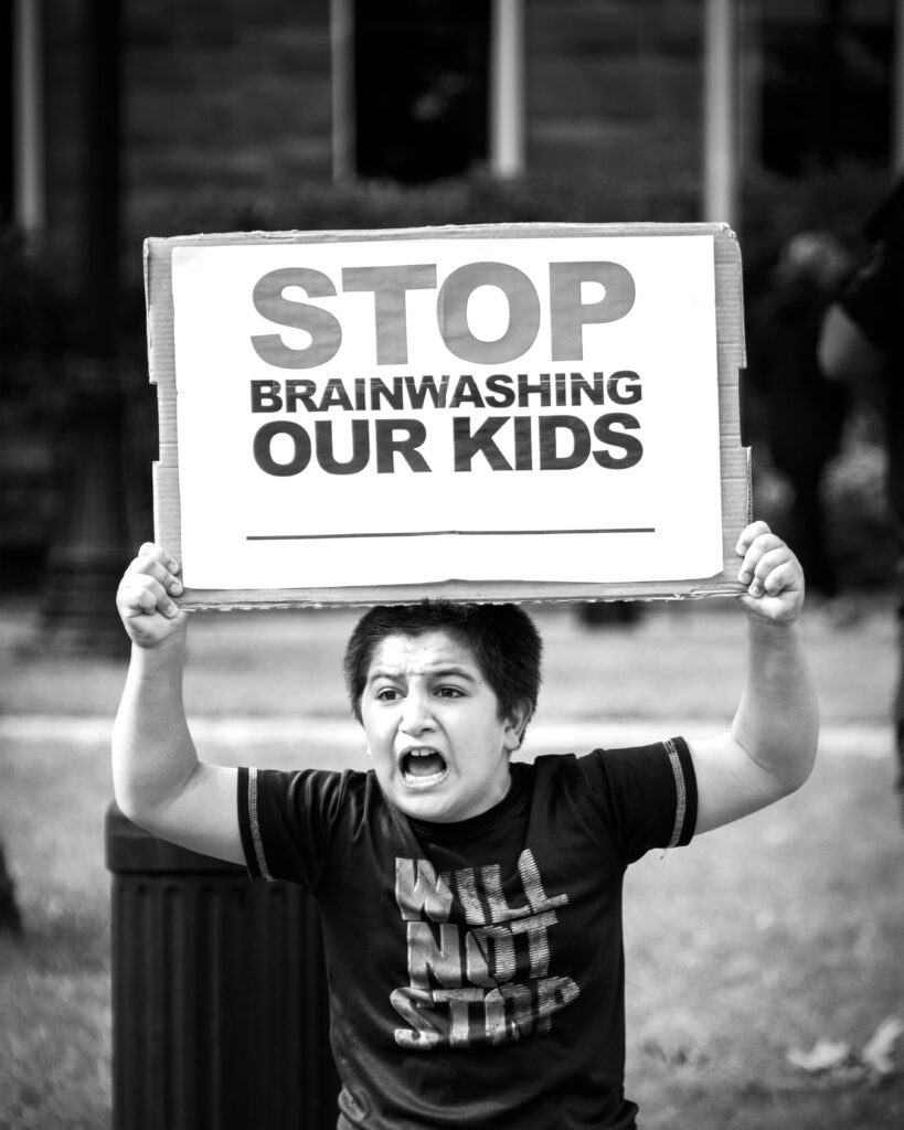 black and white image of young boy with brows furrowed and mouth open, holding a sign which says "STOP BRAINWASHING OUR KIDS"