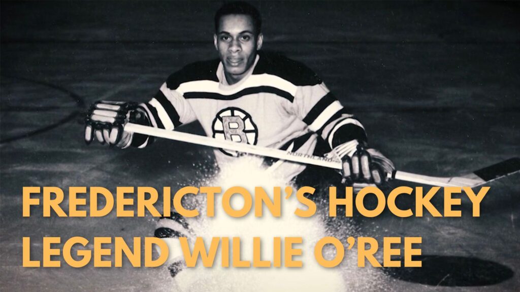 yellow text "FREDERICTON"S HOCKEY LEGEND WILLIE O'REE" over black and white image of hockey player Willie O'Ree holding a hockey stick and wearing a Boston Bruins Jersey.