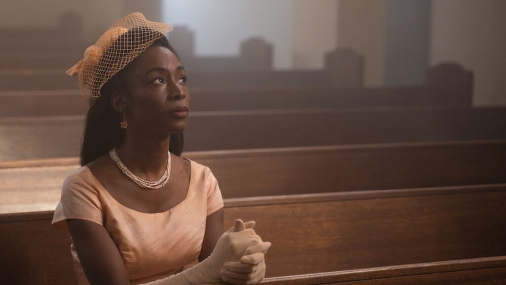 image of a well dressed Black woman in a light pink dress, gloves, and hat, sitting in church pews