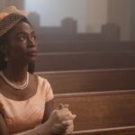 image of a well dressed Black woman in a light pink dress, gloves, and hat, sitting in church pews